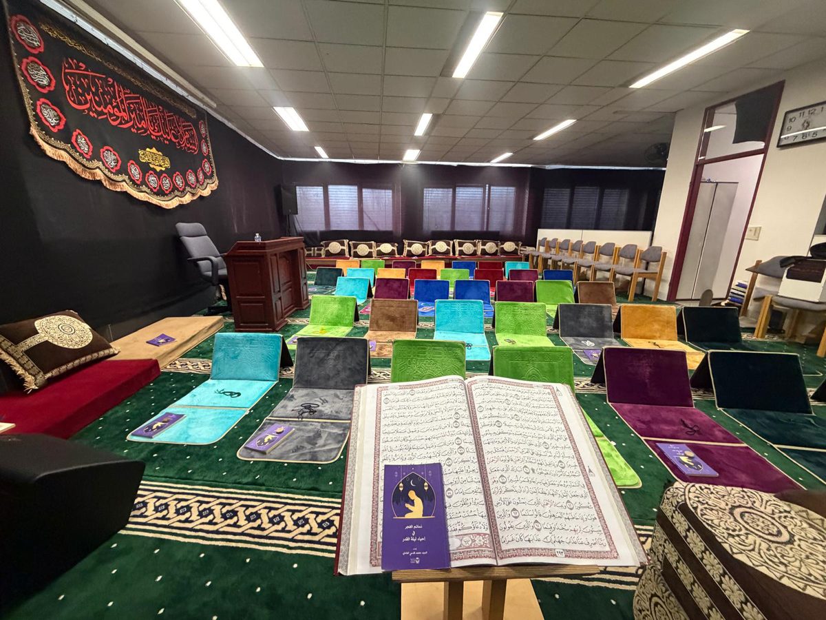 Blick in den Gebetsraum: ein aufgeschlagener Koran auf einem Holzständer, dahinter bunte Gebetsteppiche in Reihen auf grünem Teppichboden.