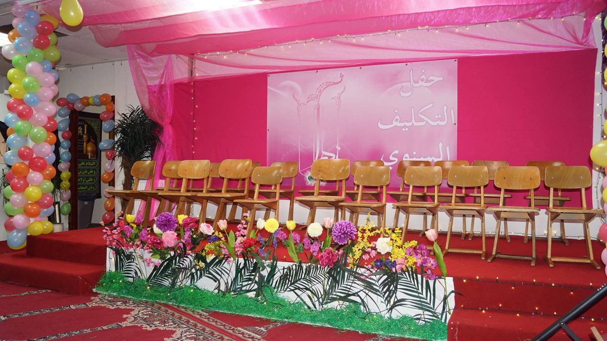 Festsaal zum jährlichen Taklif-Fest: Stuhlreihen und Blumenschmuck vor einer rosafarbenen Bühnenwand.